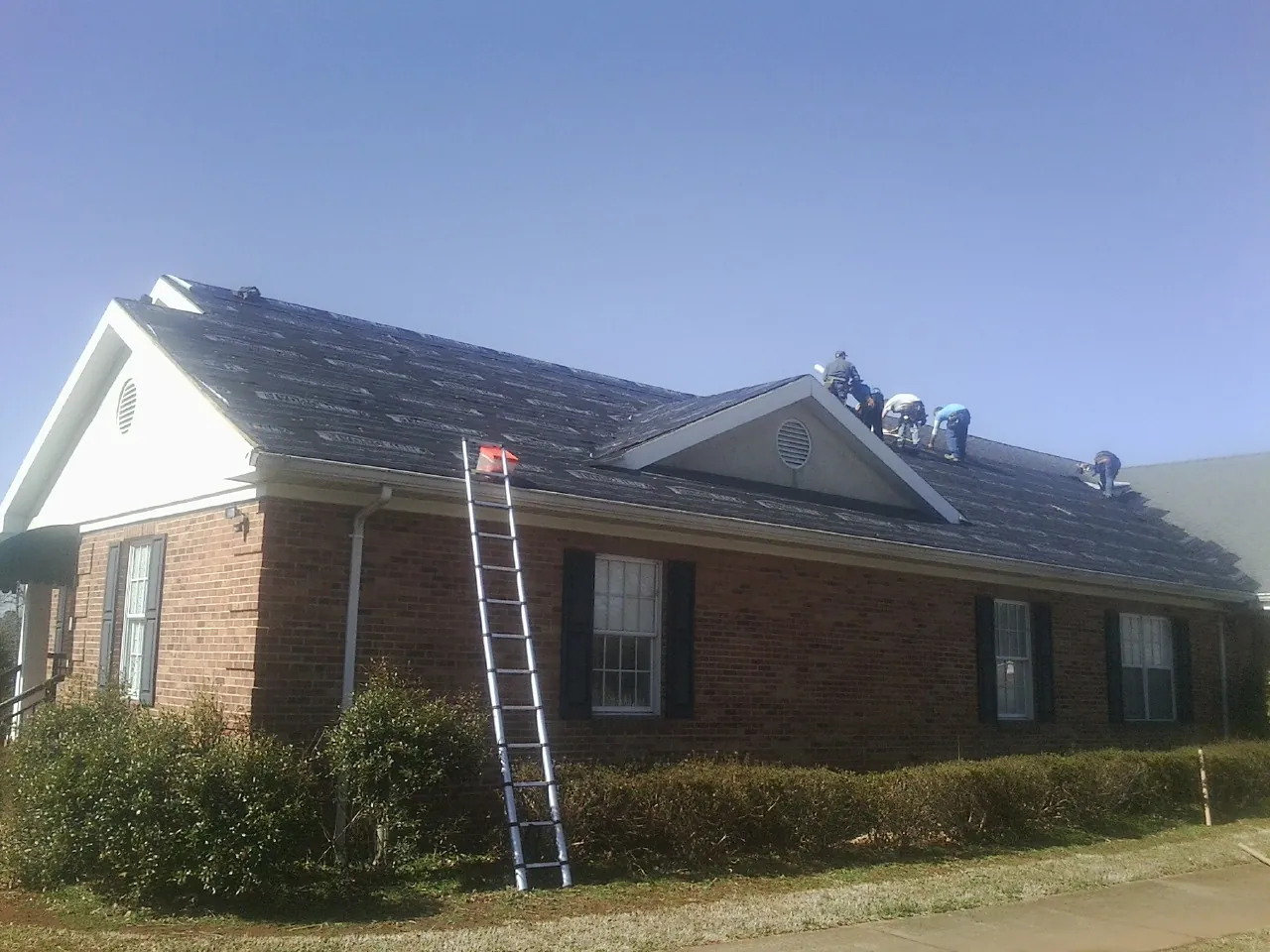 Crew tearing shingles off a brick ranch roof with a ladder set — Emergency Roof Repair work by our crew in Stedman