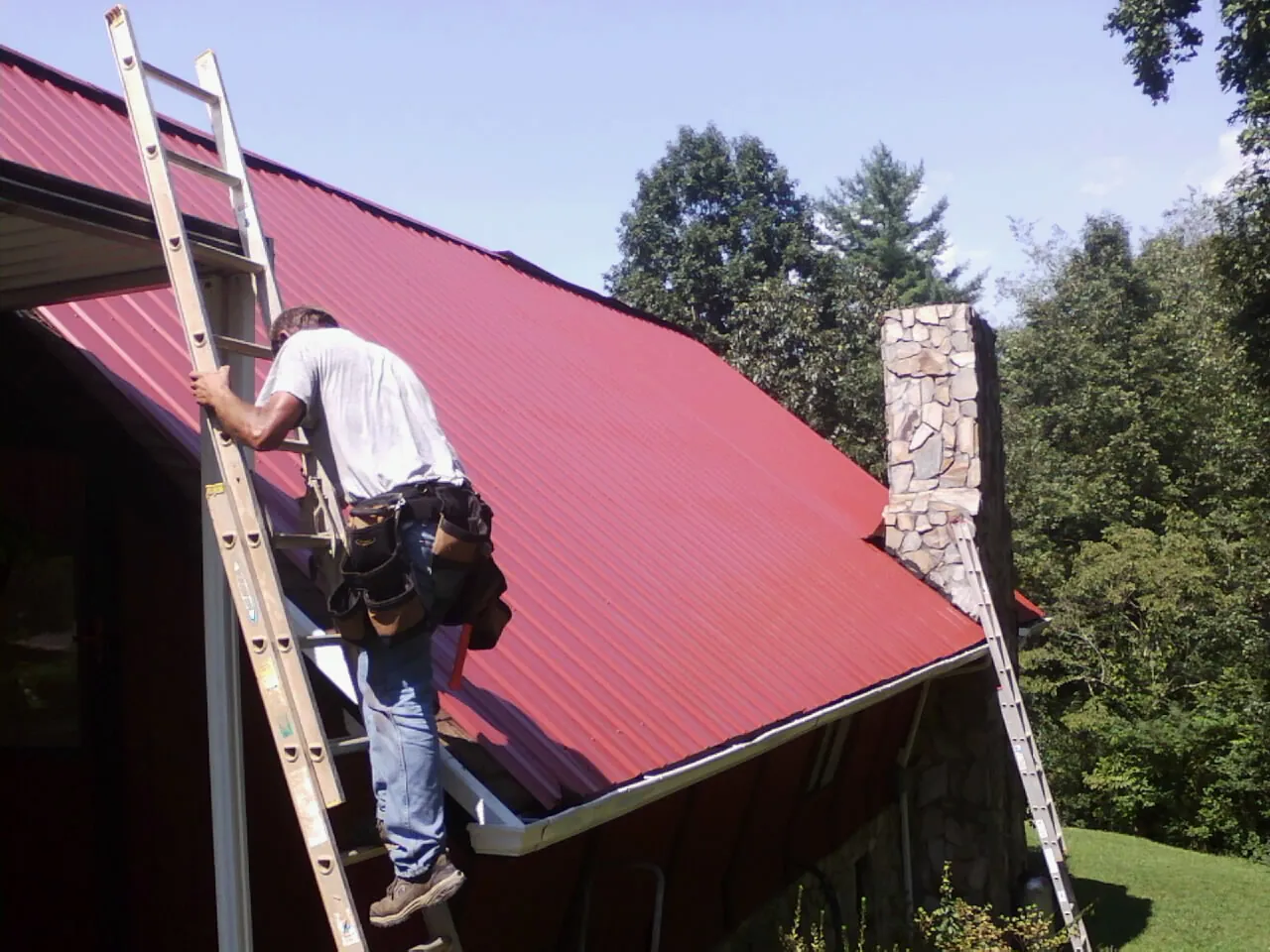 Roofer on a ladder beside a red metal roof and stone chimney — Soffit & Fascia Repair by Roofing Brothers in Stedman